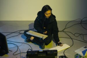 Woman in black jacket interacting with multiple keyboards on the floor in a dimly lit room.