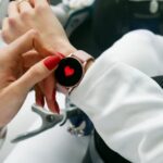 Close-up of a woman's hand with red nails interacting with a smartwatch displaying a heart icon.