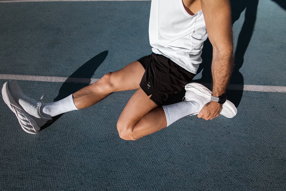 Male athlete stretching on an outdoor track, wearing white sneakers and black shorts for fitness.