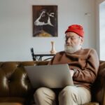 Elderly man wearing red beanie using laptop on sofa at home.