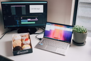 A workspace featuring a laptop, desktop monitor, box of donuts, and plant on a table.